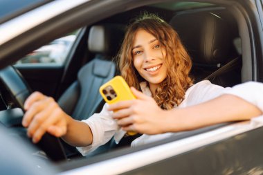 Kıvırcık saçlı ve telefonu olan genç bir kadın arabanın direksiyonunda oturur ve güneşli bir günde mesajlaşır. Güzel bir kadın elinde telefonla gezmekten zevk alır. Kentsel ulaşım kavramı.