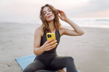 Mavi minderli, telefonlu, kablosuz kulaklıklı bir kadın denizin arka planında. Atletik kadın müzik dinler ve deniz manzarasının önünde gün doğumundan zevk alır. Meditasyon ve rahatlama kavramı.