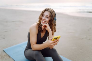 Mavi minderli, telefonlu, kablosuz kulaklıklı bir kadın denizin arka planında. Atletik kadın müzik dinler ve deniz manzarasının önünde gün doğumundan zevk alır. Meditasyon ve rahatlama kavramı.