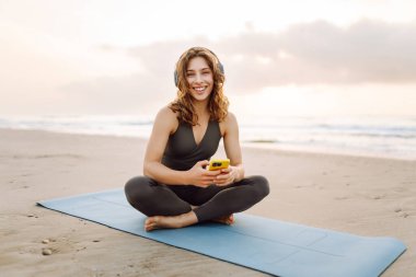 Mavi minderli, telefonlu, kablosuz kulaklıklı bir kadın denizin arka planında. Atletik kadın müzik dinler ve deniz manzarasının önünde gün doğumundan zevk alır. Meditasyon ve rahatlama kavramı.