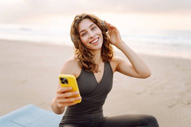Mavi minderli, telefonlu, kablosuz kulaklıklı bir kadın denizin arka planında. Atletik kadın müzik dinler ve deniz manzarasının önünde gün doğumundan zevk alır. Meditasyon ve rahatlama kavramı.