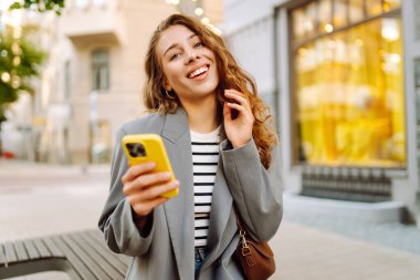 Güneşli bir günde şehir sokaklarında telefonu olan şık bir kadının portresi. Telefon mesajları atan ve gün batımında yürüyüş yapmaktan hoşlanan güzel bir kadın. Eğlence ve rahatlama kavramı.