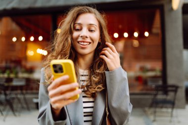 Güneşli bir günde şehir sokaklarında telefonu olan şık bir kadının portresi. Telefon mesajları atan ve gün batımında yürüyüş yapmaktan hoşlanan güzel bir kadın. Eğlence ve rahatlama kavramı.