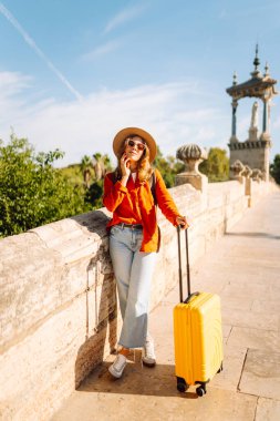 Şapkalı mutlu bir kadın, parlak sarı bir bavul taşıyor ve güneşli bir köprüde cep telefonu tutuyor. Bir turist gün batımından zevk alır, mesajlaşır. Turizm ve eğlence kavramı.