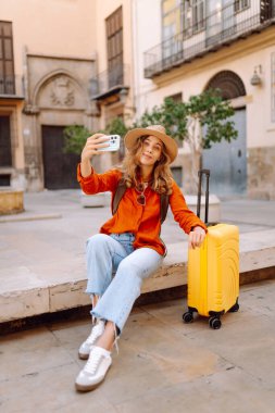 Gözlüklü, parlak renkli bavul ve telefon taşıyan güzel bir kadın şehir meydanında dolaşmaktan hoşlanıyor. Şehrin güneşli sokaklarında sırt çantalı ve bavul bloglu bir turist. Günlük konseptleri.