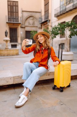Gözlüklü, parlak renkli bavul ve telefon taşıyan güzel bir kadın şehir meydanında dolaşmaktan hoşlanıyor. Şehrin güneşli sokaklarında sırt çantalı ve bavul bloglu bir turist. Günlük konseptleri.