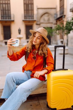 Gözlüklü, parlak renkli bavul ve telefon taşıyan güzel bir kadın şehir meydanında dolaşmaktan hoşlanıyor. Şehrin güneşli sokaklarında sırt çantalı ve bavul bloglu bir turist. Günlük konseptleri.