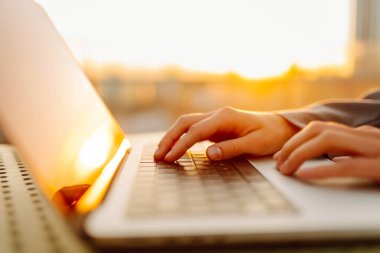 Günbatımında dizüstü bilgisayarda daktilo eden modern bir kadının yakın çekimi. Dişi bir serbest çalışan, dışarıda bir terasta dizüstü bilgisayarda çalışıyor. İş ve teknoloji kavramı.