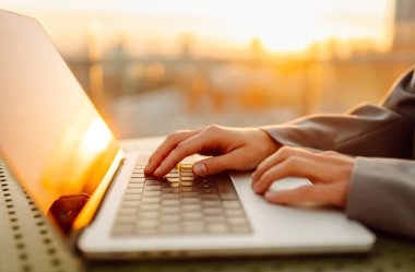 Günbatımında dizüstü bilgisayarda daktilo eden modern bir kadının yakın çekimi. Dişi bir serbest çalışan, dışarıda bir terasta dizüstü bilgisayarda çalışıyor. İş ve teknoloji kavramı.