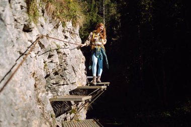 Sırt çantalı bir kadın yürüyüşçü kayalık bir ormanda yol boyunca yürür. Yürüyüşe çıkmış genç bir kadın, gün batımının ve dağlardaki maceranın tadını çıkarıyor. Aktif yaşam tarzı. Kavram: eğlence, yürüyüş.