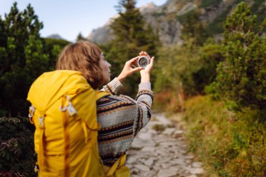 Sarı sırt çantalı ve kayalık yüzlü genç bir kadın yürüyüşçünün dikiz görüntüsü. Kadın yönünü pusula ile kontrol ediyor ve dağlarda günbatımının tadını çıkarıyor. Yön bulma ve yürüyüş kavramı.