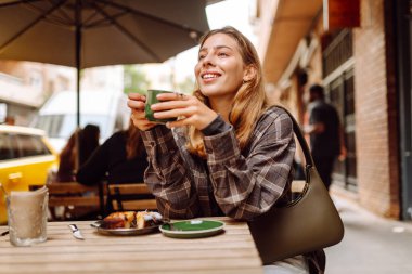 Genç bir kadın açık kafede oturmuş, içkisini yudumluyor ve telefon kullanıyor. Kadınlar güneşli bir günde mis kokulu pastalardan ve taze demlenmiş kahveden hoşlanır. Yiyecek, içecek, rahatlama, atıştırma kavramı.