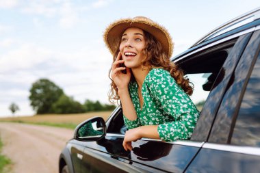 Neşeli kadın güneşli bir günde pencereden sarkmış kırsal manzaranın tadını çıkarıyor. Parlak elbiseli ve şapkalı genç gezgin, araba camından rüzgarı saçlarıyla yakalıyor. Kavram: ulaşım, macera.