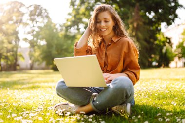 Güneşli bir günde elinde dizüstü bilgisayarla parkta çimlerin üzerinde oturan tatlı bir kadın. Dişi bir serbest yazar ya da öğrenci dışarıda bir dizüstü bilgisayarda çalışıyor. Konsept: Eğitim, uzak çalışma, açık hava.