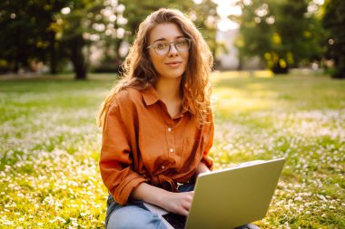 Güneşli bir günde elinde dizüstü bilgisayarla parkta çimlerin üzerinde oturan tatlı bir kadın. Dişi bir serbest yazar ya da öğrenci dışarıda bir dizüstü bilgisayarda çalışıyor. Konsept: Eğitim, uzak çalışma, açık hava.