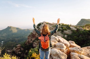 Sırt çantalı kadın yürüyüşçü gün batımında dağlarda yürüyüş yolu boyunca yürür. Güzel bir kadın dağ manzarasından zevk alır ve güneş ışınlarıyla kayaların üzerinde yürür. Konseptler: macera, yürüyüş ve doğa.