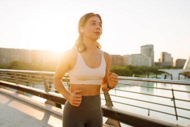 Kablosuz kulaklık takan atletik bir kadın güneş ışınlarıyla köprüden geçiyor. Güneşli bir günde şehrin tadını çıkaran atletik giysiler içindeki genç bir kadın. Koşu, spor ve sağlık kavramları.