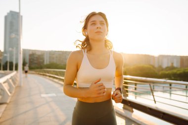 Kablosuz kulaklık takan atletik bir kadın güneş ışınlarıyla köprüden geçiyor. Güneşli bir günde şehrin tadını çıkaran atletik giysiler içindeki genç bir kadın. Koşu, spor ve sağlık kavramları.