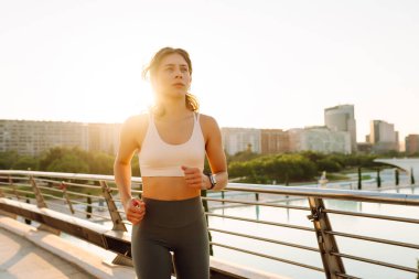 Kablosuz kulaklık takan atletik bir kadın güneş ışınlarıyla köprüden geçiyor. Güneşli bir günde şehrin tadını çıkaran atletik giysiler içindeki genç bir kadın. Koşu, spor ve sağlık kavramları.