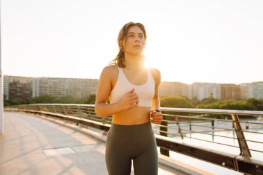 Kablosuz kulaklık takan atletik bir kadın güneş ışınlarıyla köprüden geçiyor. Güneşli bir günde şehrin tadını çıkaran atletik giysiler içindeki genç bir kadın. Koşu, spor ve sağlık kavramları.