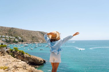 Genç bir kadın bir uçurumda oturuyor, şapkası var, deniz manzarasına bakıyor. Özgür hisseden bir kadının deniz kenarında güneşli bir günün tadını çıkarması. Doğa, özgürlük ve hafta sonları kavramları.