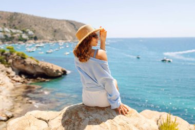 Genç bir kadın bir uçurumda oturuyor, şapkası var, deniz manzarasına bakıyor. Özgür hisseden bir kadının deniz kenarında güneşli bir günün tadını çıkarması. Doğa, özgürlük ve hafta sonları kavramları.