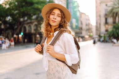 Şapkalı ve sırt çantalı güzel bir turist şehrin güneşli sokaklarında geziniyor. Mutlu bir kadın günbatımında Central Square 'in mimarisine hayran. Turizm kavramları, yürüyüş. Seyahat tutkusu.