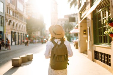 Şapkalı ve sırt çantalı güzel bir turist şehrin güneşli sokaklarında geziniyor. Mutlu bir kadın günbatımında Central Square 'in mimarisine hayran. Turizm kavramları, yürüyüş. Seyahat tutkusu.
