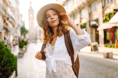 Şapkalı ve sırt çantalı güzel bir turist şehrin güneşli sokaklarında geziniyor. Mutlu bir kadın günbatımında Central Square 'in mimarisine hayran. Turizm kavramları, yürüyüş. Seyahat tutkusu.