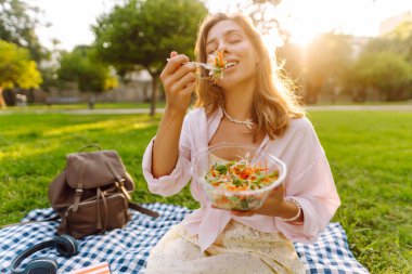 Güneşli bir parkta salata yiyen güzel bir kadının portresi. Genç bir kadın mavi bir paspasın üzerinde oturur, günbatımının tadını çıkarır ve dışarıda atıştırır. Konseptler: yiyecek, kentsel ve boş zaman.