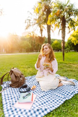 Güneşli bir parkta salata yiyen güzel bir kadının portresi. Genç bir kadın mavi bir paspasın üzerinde oturur, günbatımının tadını çıkarır ve dışarıda atıştırır. Konseptler: yiyecek, kentsel ve boş zaman.