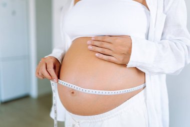 Hamile bir kadın, göbeğini parlak bir odada mezurayla ölçer. Genç bir kadın, bebeğinin rahimde büyümesini izliyor. Odak noktası sağlık, kontrol ve rahatlık..