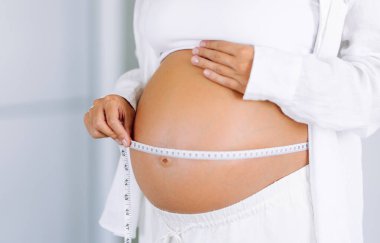 Hamile bir kadın, göbeğini parlak bir odada mezurayla ölçer. Genç bir kadın, bebeğinin rahimde büyümesini izliyor. Odak noktası sağlık, kontrol ve rahatlık..