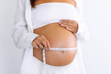 Hamile bir kadın, göbeğini parlak bir odada mezurayla ölçer. Genç bir kadın, bebeğinin rahimde büyümesini izliyor. Odak noktası sağlık, kontrol ve rahatlık..