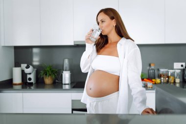 Hamile bir kadın modern bir mutfakta duruyor ve sıcak su içiyor. Çocuk bekleyen genç bir kadın sıcak bir odada güneşli bir günün tadını çıkarıyor. Sağlık ve içecek kavramı.