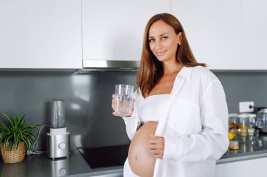 Hamile bir kadın modern bir mutfakta duruyor ve sıcak su içiyor. Çocuk bekleyen genç bir kadın sıcak bir odada güneşli bir günün tadını çıkarıyor. Sağlık ve içecek kavramı.