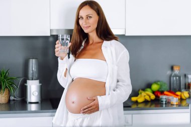 Hamile bir kadın modern bir mutfakta duruyor ve sıcak su içiyor. Çocuk bekleyen genç bir kadın sıcak bir odada güneşli bir günün tadını çıkarıyor. Sağlık ve içecek kavramı.