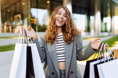 Modern bir binanın önünde alışveriş torbalarıyla şık kıyafetler içinde neşeli bir kadın. Şık bir kadın alışverişten sonra güneşli bir günün tadını çıkarır. Tüketim kavramı, alışveriş.