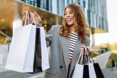 Modern bir binanın önünde alışveriş torbalarıyla şık kıyafetler içinde neşeli bir kadın. Şık bir kadın alışverişten sonra güneşli bir günün tadını çıkarır. Tüketim kavramı, alışveriş.