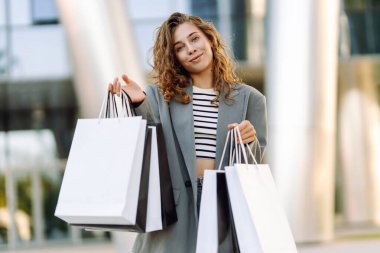 Modern bir binanın önünde alışveriş torbalarıyla şık kıyafetler içinde neşeli bir kadın. Şık bir kadın alışverişten sonra güneşli bir günün tadını çıkarır. Tüketim kavramı, alışveriş.
