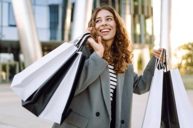 Modern bir binanın önünde alışveriş torbalarıyla şık kıyafetler içinde neşeli bir kadın. Şık bir kadın alışverişten sonra güneşli bir günün tadını çıkarır. Tüketim kavramı, alışveriş.