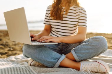 Dizüstü bilgisayarı olan genç bir kadın güneşin altında bir plaj paspasında oturuyor. Gözlüklü bir kadın serbest çalışıyor ve deniz manzarasının tadını çıkarıyor. Uzaktan çalışma ve rahatlama kavramı.