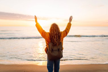 Sırt çantalı gülümseyen bir kadın kumsalda günbatımının tadını çıkarıyor. Bir yürüyüşçü gün doğumunda kumlu bir plaj boyunca yürür. Yürüyüş, doğa ve özgürlük kavramı.