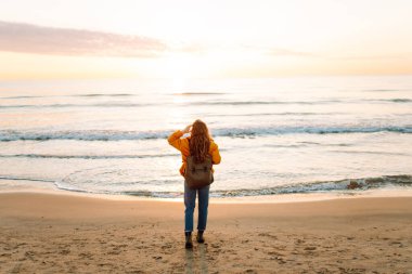 Sırt çantalı gülümseyen bir kadın kumsalda günbatımının tadını çıkarıyor. Bir yürüyüşçü gün doğumunda kumlu bir plaj boyunca yürür. Yürüyüş, doğa ve özgürlük kavramı.
