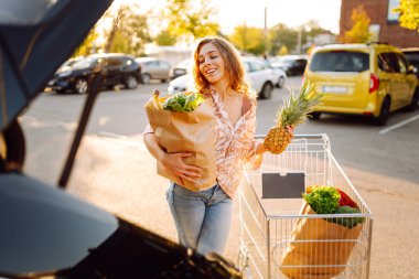 Park yerinde arabasının yanında taze yiyeceklerle duran genç bir kadının portresi. Güzel bir kadın arabanın bagajına meyve ve sebze koyar. Alışveriş, tüketim ve kentsel yaşam kavramları.