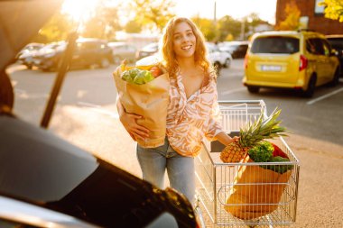 Park yerinde arabasının yanında taze yiyeceklerle duran genç bir kadının portresi. Güzel bir kadın arabanın bagajına meyve ve sebze koyar. Alışveriş, tüketim ve kentsel yaşam kavramları.