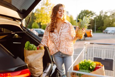 Park yerinde arabasının yanında taze yiyeceklerle duran genç bir kadının portresi. Güzel bir kadın arabanın bagajına meyve ve sebze koyar. Alışveriş, tüketim ve kentsel yaşam kavramları.