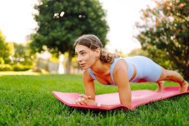 Atletik giysiler içinde güzel bir kadın güneşli bir parkta yoga minderinde antrenman yapıyor. Atletik bir kadın dışarıda çeşitli egzersizler yapar. Spor, rahatlama ve aktif bir yaşam tarzı kavramları.