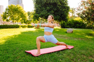 Atletik giysiler içinde güzel bir kadın güneşli bir parkta yoga minderinde antrenman yapıyor. Atletik bir kadın dışarıda çeşitli egzersizler yapar. Spor, rahatlama ve aktif bir yaşam tarzı kavramları.
