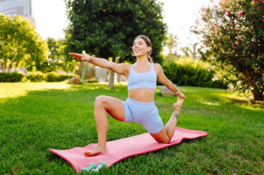 Atletik giysiler içinde güzel bir kadın güneşli bir parkta yoga minderinde antrenman yapıyor. Atletik bir kadın dışarıda çeşitli egzersizler yapar. Spor, rahatlama ve aktif bir yaşam tarzı kavramları.